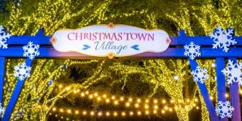 A sign saying "Christmas Town Village" on an archway in front of trees lit with strings of lights
