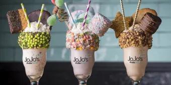 Three large milkshakes in a row topped with whipped cream, sprinkles, marshmallows and cookies