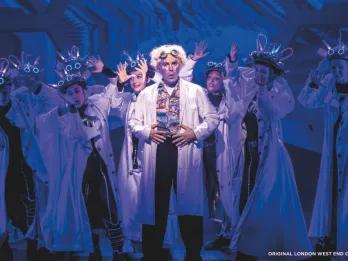 Back to the Future The Musical Doc Brown wearing a white lab coat standing centre stage in front of dancers also wearing lab coats