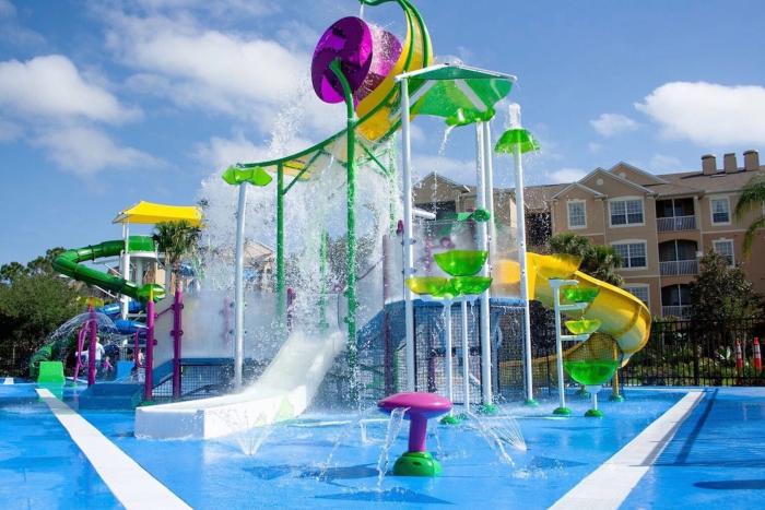A colourful children's water splash pad playground with water fountains, slides, and a dumping wheel under a blue sky.