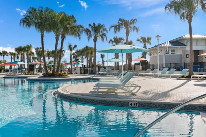 Resort swimming pool lined with lounge chairs, umbrellas, and palm trees under a bright blue sky.