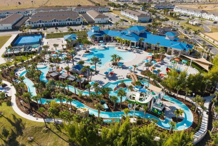 An aerial drone view of a resort water park featuring a large swimming pool, lazy river, water slides, sports courts, and a main clubhouse building with a blue roof.