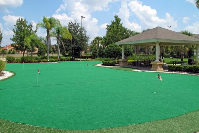 An outdoor resort putting green with golf flags next to a shaded picnic pavilion with tables under a cloudy blue sky.