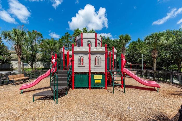 A grey and red castle-themed children's playground structure with slides on a wood-chip surface under a blue sky.