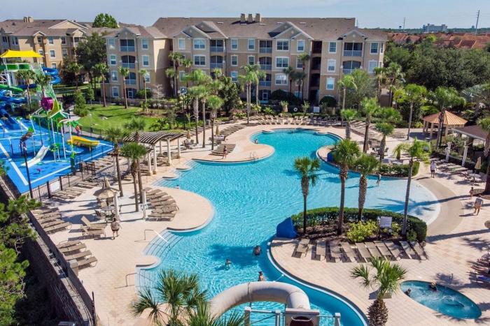 Aerial overview of a lagoon-style resort swimming pool, water slides, and multi-story vacation condo buildings under a clear sky.