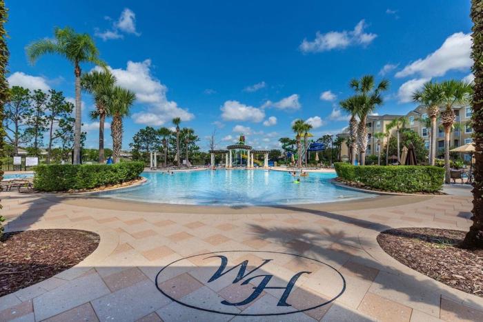 Ground-level view of a resort pool deck with a stone inlaid WH logo looking toward a large blue swimming pool and background water slides.