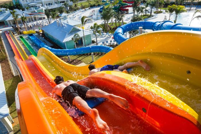 Two people racing face-first on blue mats down twisting orange and yellow resort water slides.