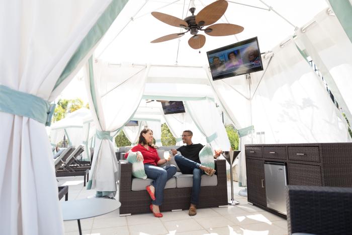 A couple relaxing on a couch inside a luxury poolside cabana with white curtains, a ceiling fan, a TV, and a mini-fridge.