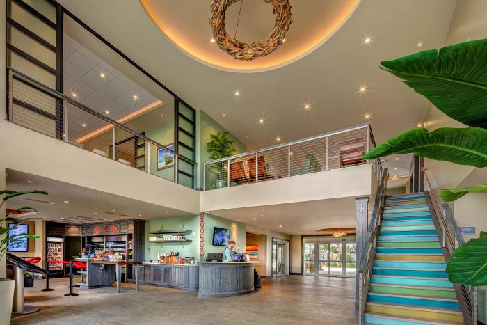 The modern, two-story lobby of a resort clubhouse featuring a concierge desk, market cafe, large stairs, and a circular ceiling fixture.