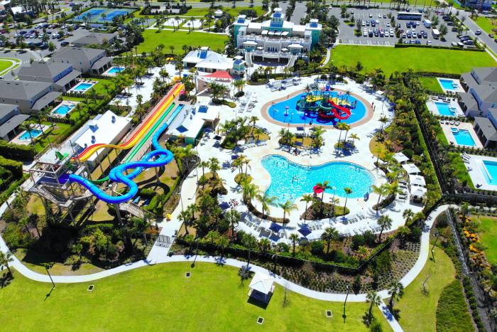 An aerial drone view of a resort water park featuring a large pool, twisting water slides, splash pad, and a main clubhouse building.