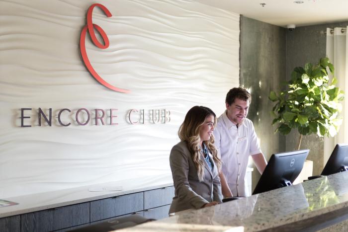 Two resort reception staff members standing at a granite service desk beneath a large Encore Club wall sign.