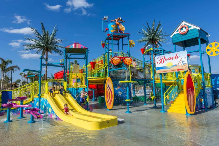 A colorful children's water playground with yellow slides, a beach sign, surfboards, and palm trees under a bright blue sky.