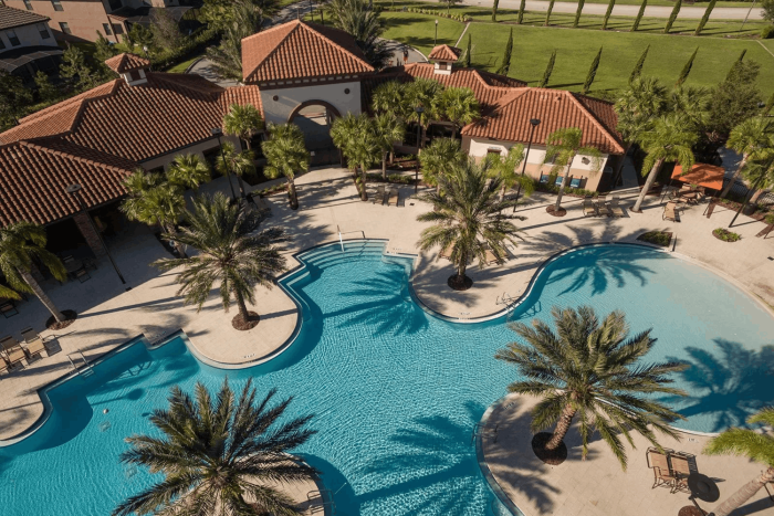 High-angle view of a curvy resort swimming pool surrounded by tall palm trees and a Spanish-style clubhouse.
