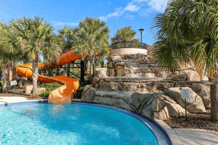 Large orange twisting water slide and a rock waterfall feature flowing into a resort swimming pool surrounded by palm trees.