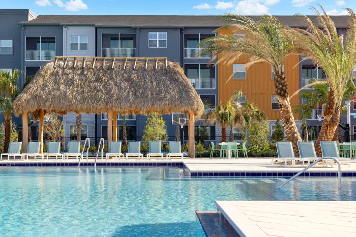 Large swimming pool lined with lounge chairs beneath a large thatched roof tiki cabana