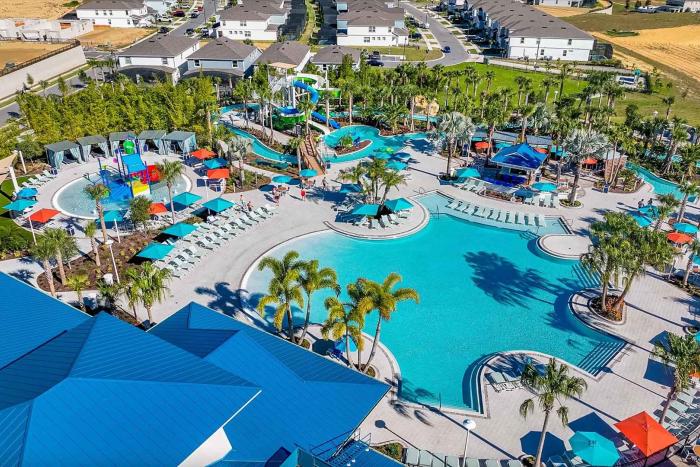 An aerial view of a large zero-entry resort pool, a children's water splash pad, water slides, and palm trees next to a bright blue roofed building.