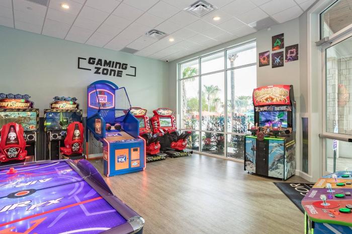 An indoor resort arcade room labeled Gaming Zone with an air hockey table, basketball arcade game, racing games, and large windows.
