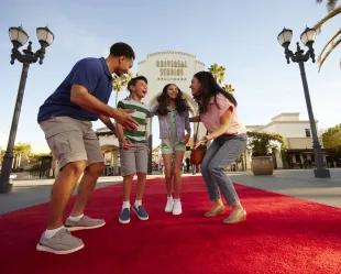 Family jumping on the red carpet in front of the large white stone entrance sign with Universal Studios Hollywood logo on. The family has dad on the right, one girl and one boy in the middle jumping and the mum crouched on the right.