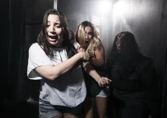 Two young women in a dark room with a bright light shining behind with terrified facial expressions, clutching onto one another and dodging another person who is lurking in the dark. The third person has black hair and a wound on their head which seems to be bleeding down their face. Their hair is messy and they are pulling a scary face with their tongue poking out.