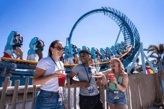 Three young adults stood in front of the Pipeline rollercoaster drinking cocktails and eating festival plates