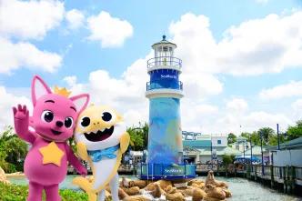 Two mascot animals, one pink dog creature wearing a yellow crown. They are stood in front of a blue lighthouse on the rocks.
