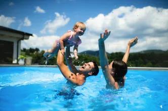 A family enjoying a high-energy "darecation" moment in a sunny outdoor pool, with water splashing as a father lifts a young child