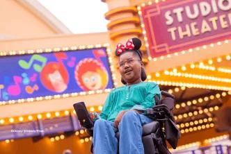 Guest in a wheelchair wearing Minnie Mouse ears smiling beneath the Studio Theater marquee at Production Courtyard, Disney Adventure World