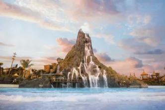 A wide shot of a massive artificial volcano with cascading waterfalls flowing into a large, wavy blue pool, under a pastel pink and blue sunset sky with scattered clouds