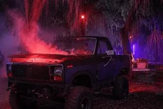 A dark pickup truck with red smoke rising from the open hood parked in swampy woods filled with neon lighting