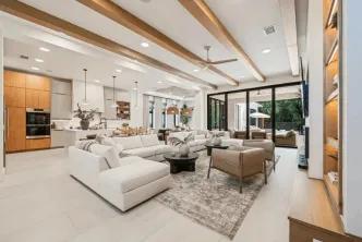 A spacious modern open-concept living room and kitchen featuring wooden ceiling beams, a large white sectional sofa, and floor-to-ceiling glass patio doors