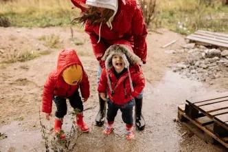 Two young children in red raincoats laugh and play in a muddy puddle while an adult in a red coat bends down beside them, illustrating a rainy day out with the family