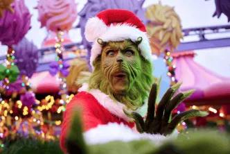 The Grinch, wearing a red Santa suit and hat, poses with a surprised expression in a brightly decorated theme park area