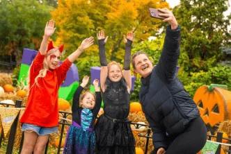 A woman taking a selfie with three young girls dressed in Halloween costumes, with pumpkins and hay bales in the background