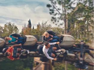 Four guests turned to the side on a coaster, sat in carts shaped like a motorbike, zooming through trees in front of the brown Hogwarts castle