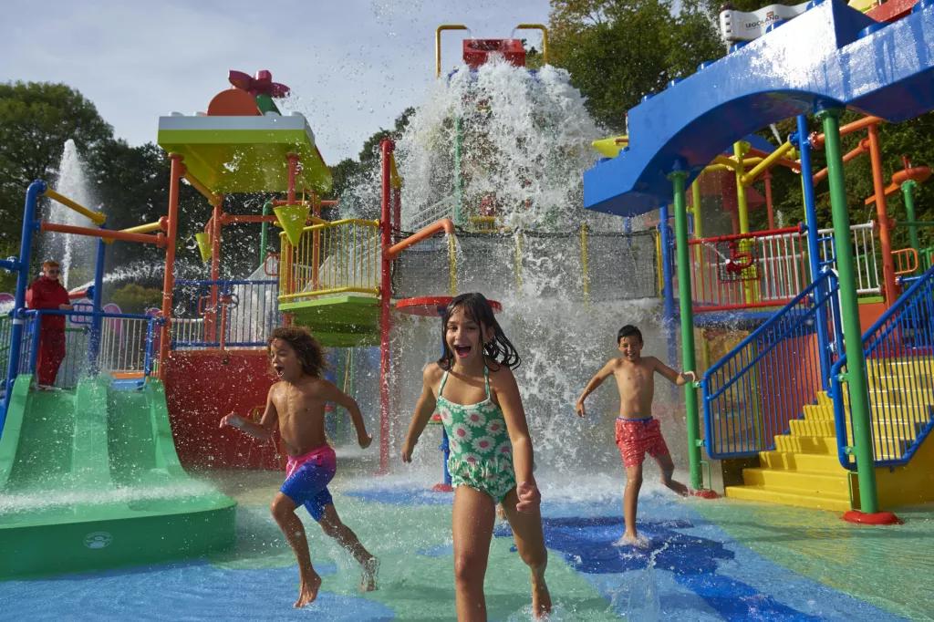 Drench Towers Water Play Area Three children in swimming costumes running away from a tipping bucket of water