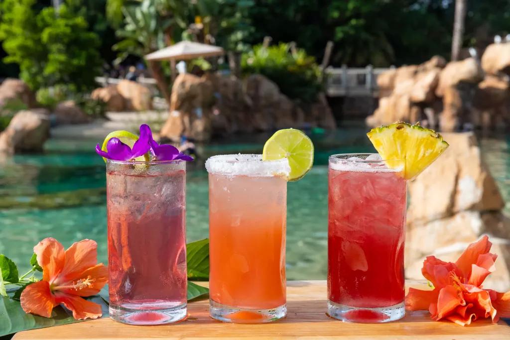 Hibiscus Hideaway Cocktails Three fruity cocktails surrounded by flowers in front of a beach