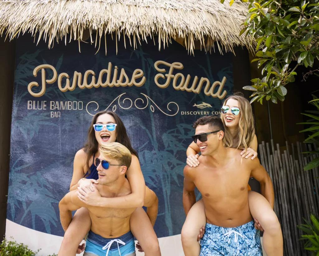 Paradise Found Wall at Blue Bamboo Bar Two men in swimming trunks giving two women piggy backs in front of a wall that says "Paradise Found"