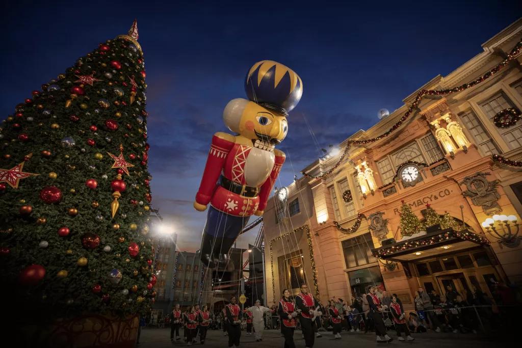 Universal's Holiday Parade featuring Macy's A large balloon shaped like a nutcracker being carried on string, passing next to a large Christmas tree