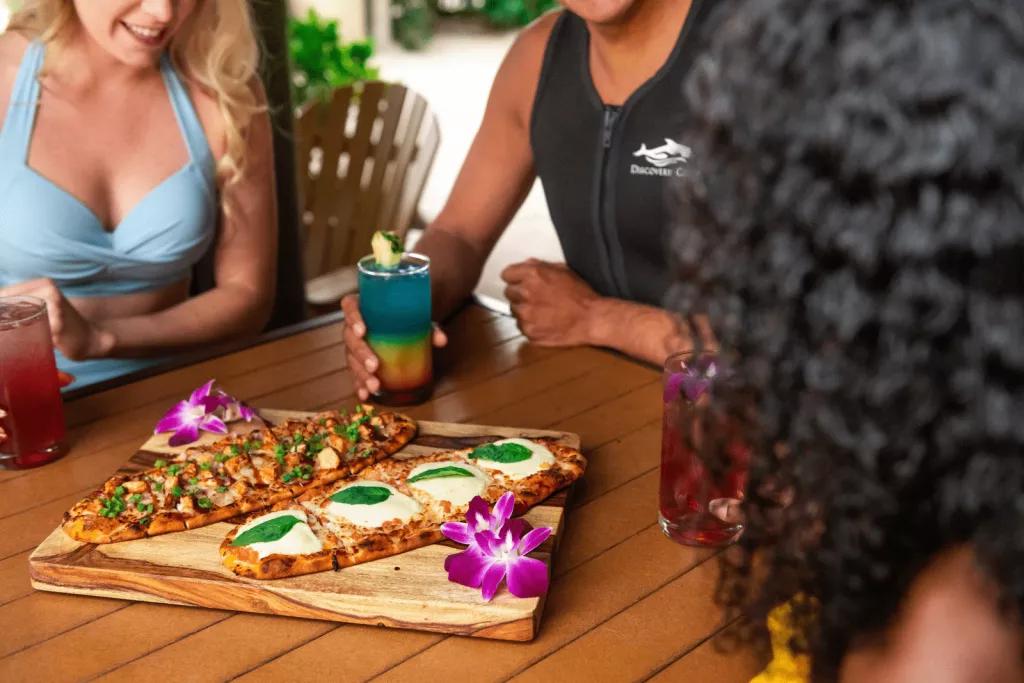 All-Inclusive Dining at Discovery Cove Three people in wetsuits and swimming costumes sitting around a wooden table with two pizzas on it