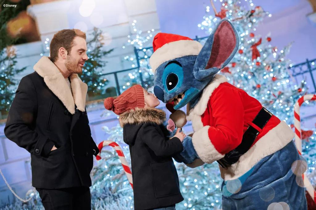 Festive Character Encounters Stitch dressed as Santa being kissed on the nose by a small child. Christmas trees and candy canes are in the background
