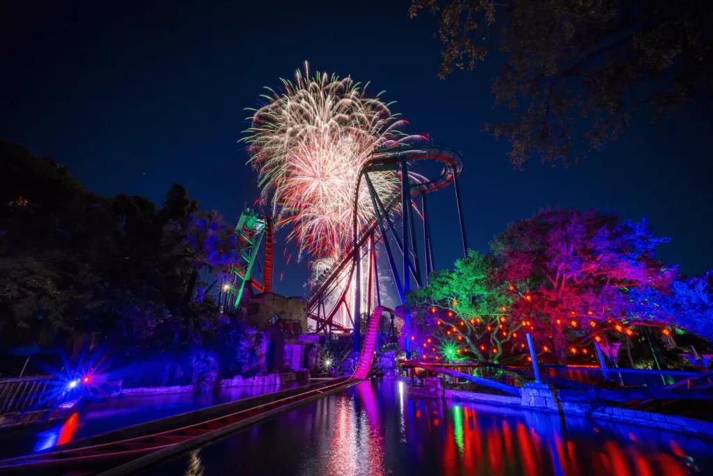 Holiday in the Sky Large colourful fireworks in a dark sky, behind a tall rollercoaster and some trees