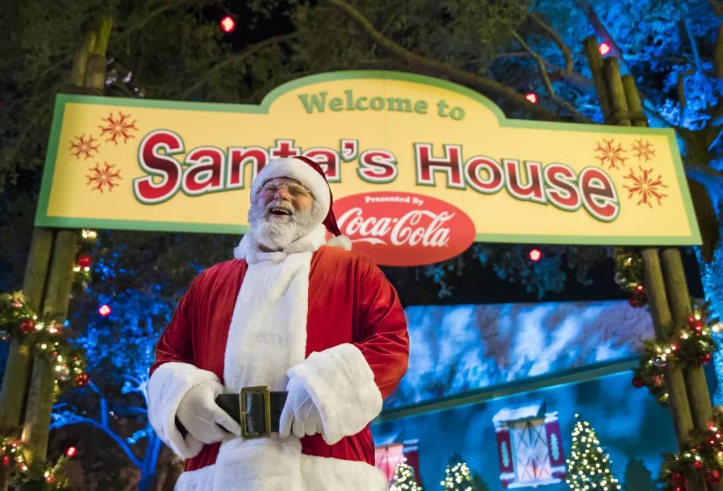 Santa's North Pole Experience Santa Clause laughing while standing in front of a sign that says "Welcome to Santa's House"
