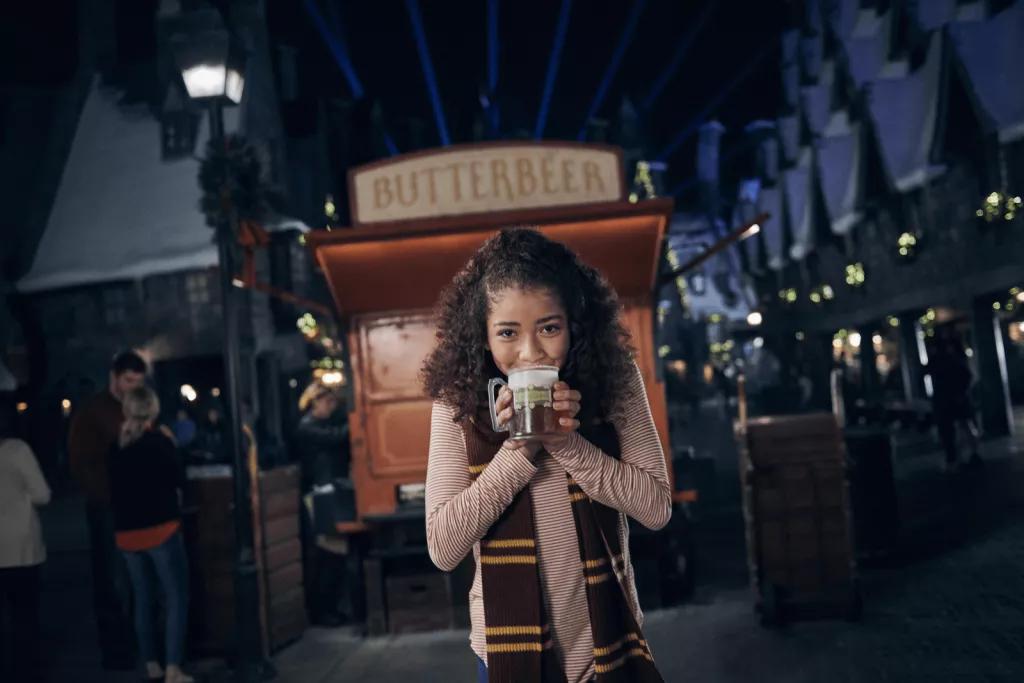 Hot Butterbeer at Wizarding World of Harry Potter A girl wearing a red a yellow scarf drinking a hot butterbeer in front of a Butterbeer cart