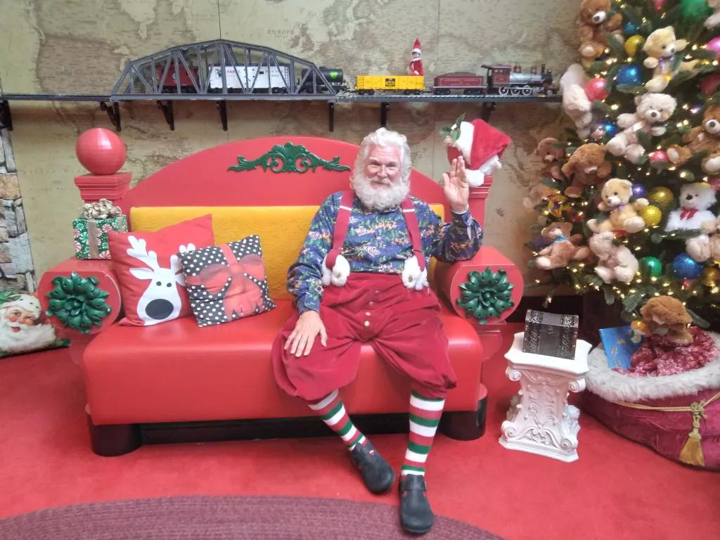 Santa Workshop Experience at ICON Park Santa Claus waving at the camera while sat on a sofa next to a Christmas tree and Christmas presents