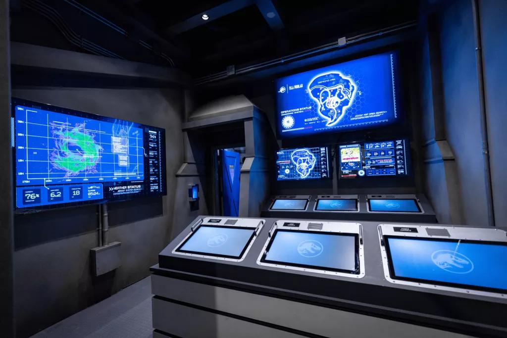 Jurassic World Escape A dark room filled with screens on countertops and on the walls showing statistics and other information