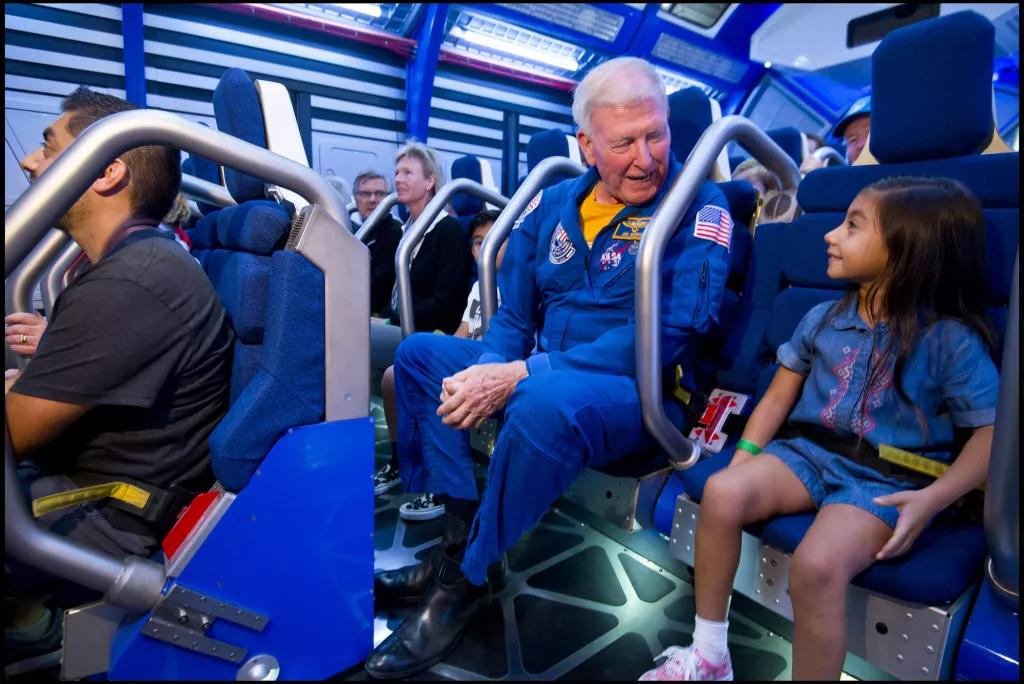 Shuttle Launch Experience An astronaut wearing a blue jumpsuit sat next to a young girl, both strapped into a simulator