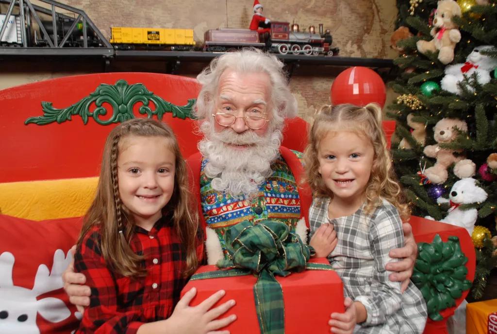 The Santa Workshop Experience Two young girls posing with Santa and a present next to a Christmas Tree