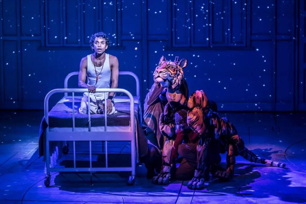 Life of Pi on Broadway A man on a stage kneeling on a single metal-framed bed next to a life-sized puppet of a tiger