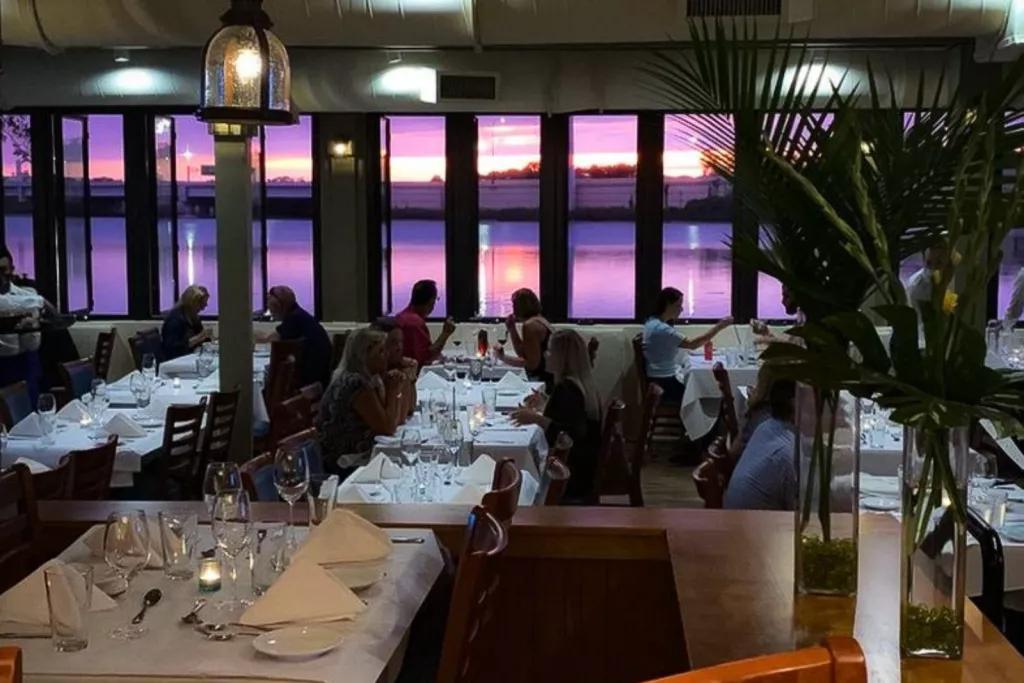 Russel's on Lake Ivanhoe A restaurant dining room with people eating at tables covered with white table cloths. There is a sunset outside reflected on the water, turning it a light purple.