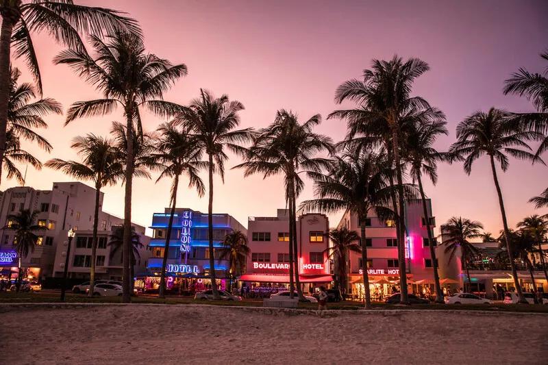 Miami at Night A row of art deco style buildings lit up with neon lights behind a sandy beach lines with tall palm trees